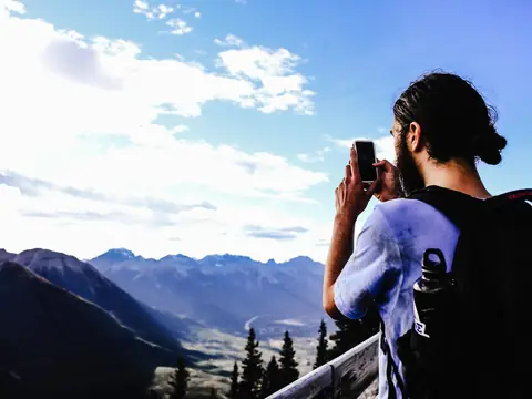 A man photographing a mountain valley on his phone under a blue sky