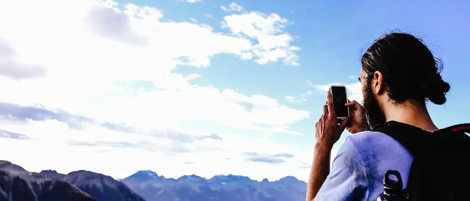 A man photographing a mountain valley on his phone under a blue sky