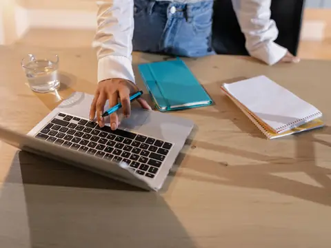 Hands on a laptop at a wooden desk with notebooks beside it