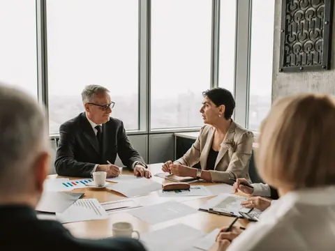 People talking over papers at a meeting table by tall windows