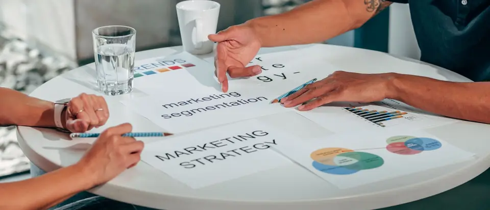 Two people pointing at a printed marketing plan on a round table