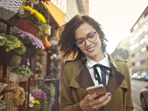 A woman smiling at her phone outside a flower shop on a sunny street