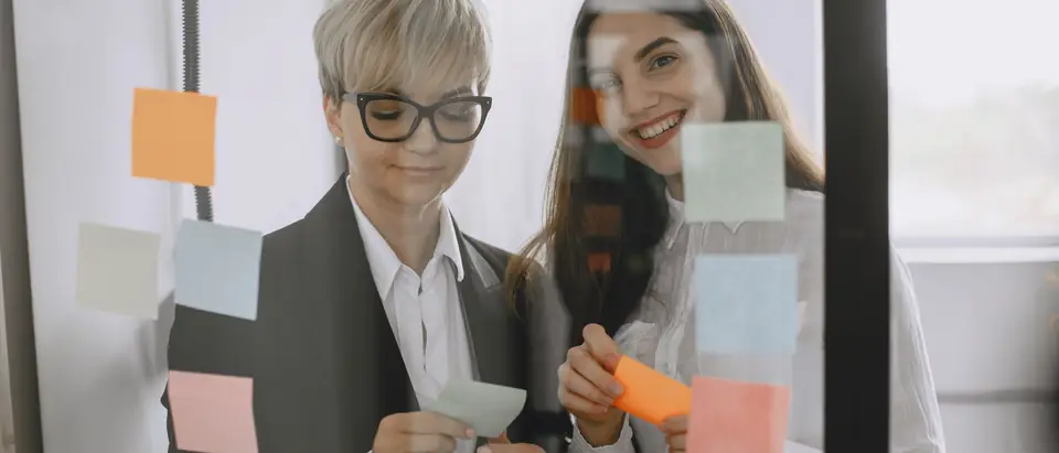 Two women sorting sticky notes on a glass wall
